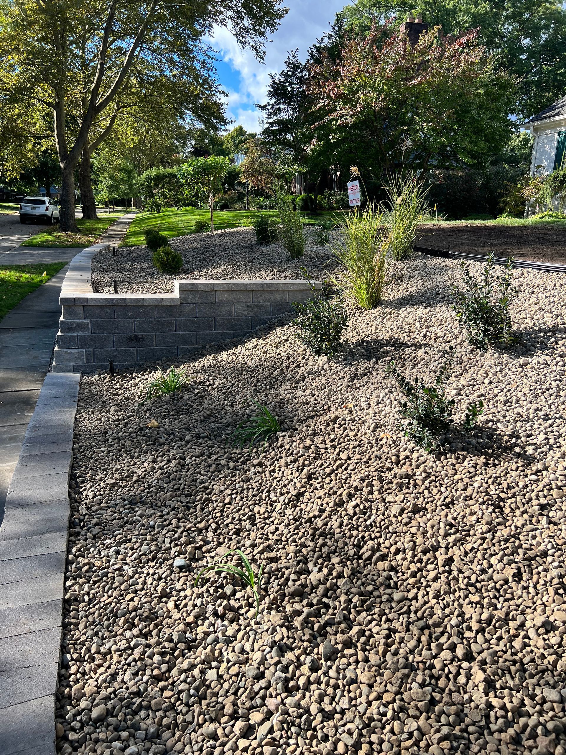 A tiered stone retaining wall borders a sloped garden bed filled with grey gravel and young shrubs on a sunny day.