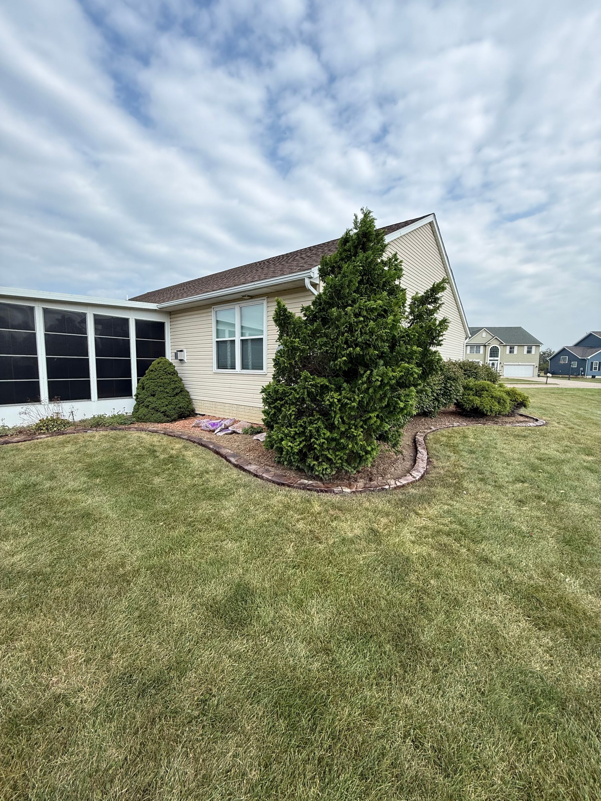 A beige house with a sunroom and landscaped shrubs in a grassy yard under a cloudy blue sky.