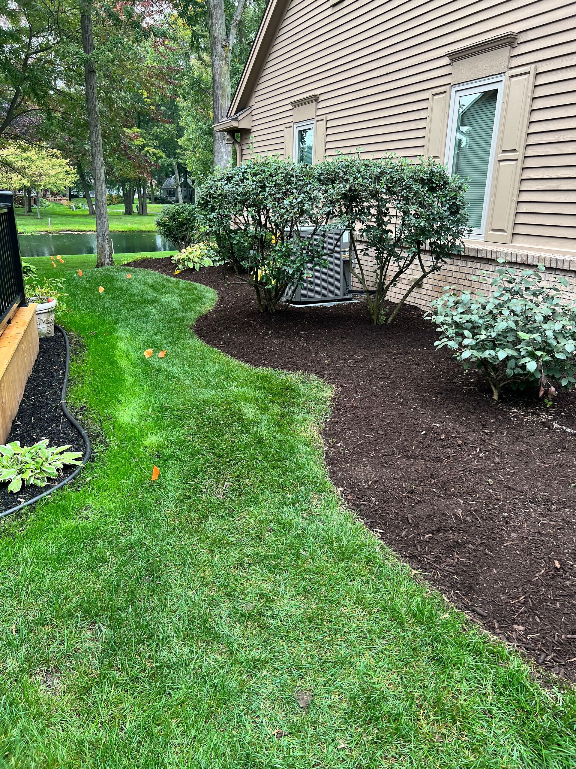 A curving grassy lawn borders a garden bed with dark mulch, green shrubs, and an HVAC unit beside a tan house exterior.
