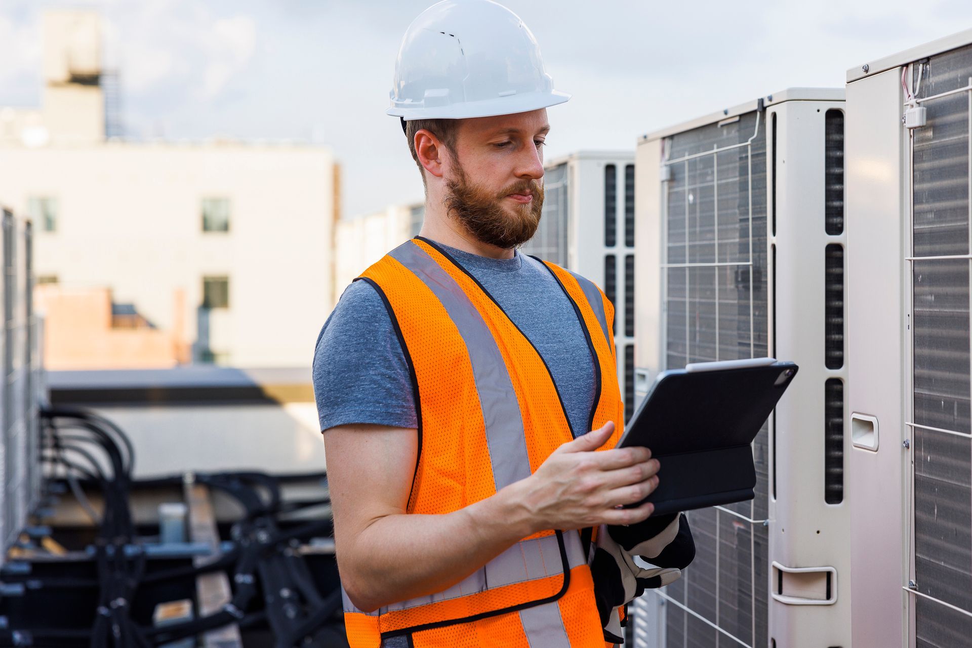 HVAC contractor in safety gear inspecting rooftop AC units with a tablet.