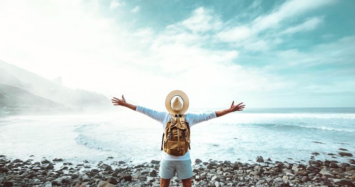 Person with arms outstretched, wearing a hat and backpack, facing the ocean. Blue sky and waves.