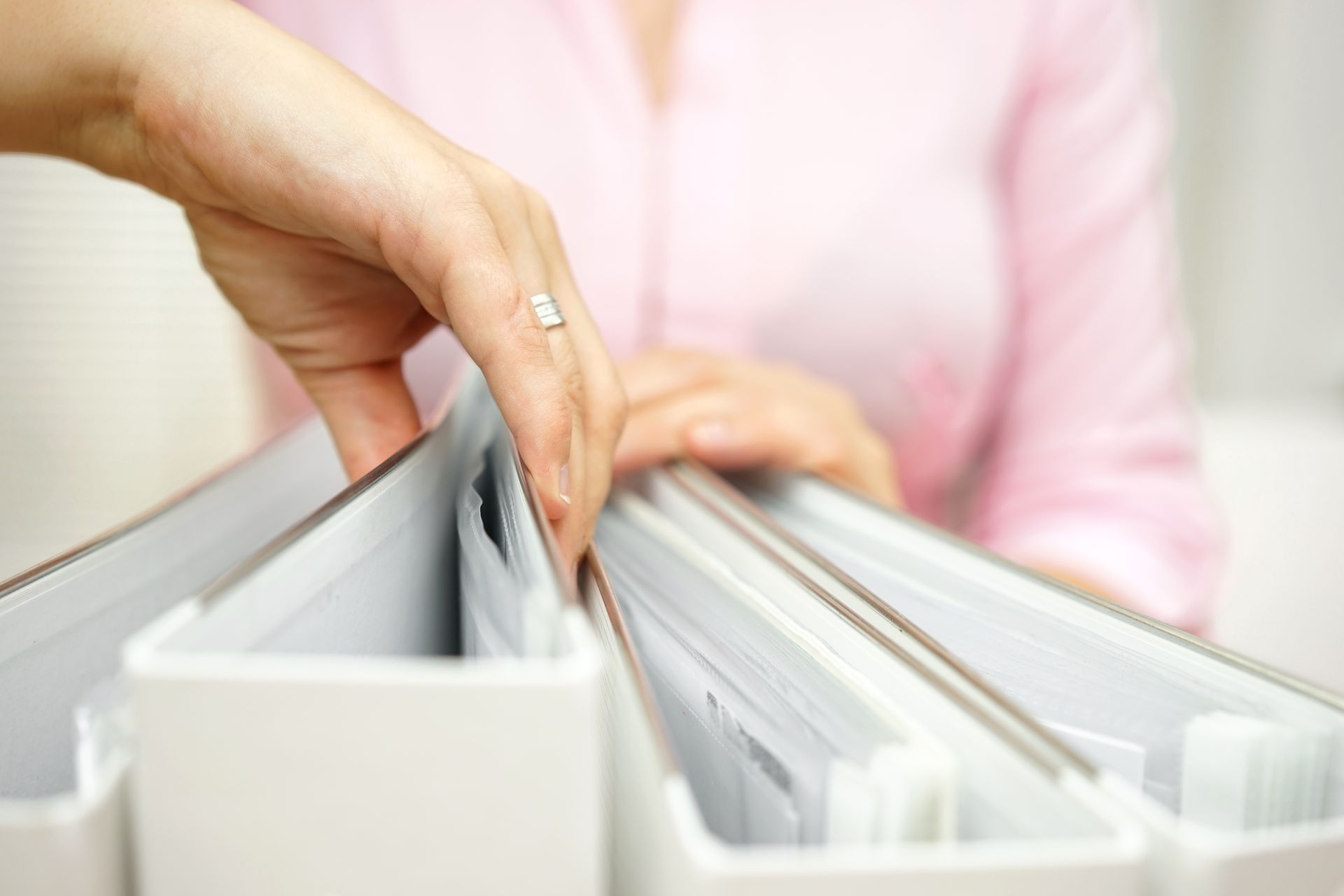 Hand reaching through white file folders in a drawer, with a blurred person in a pink shirt behind them