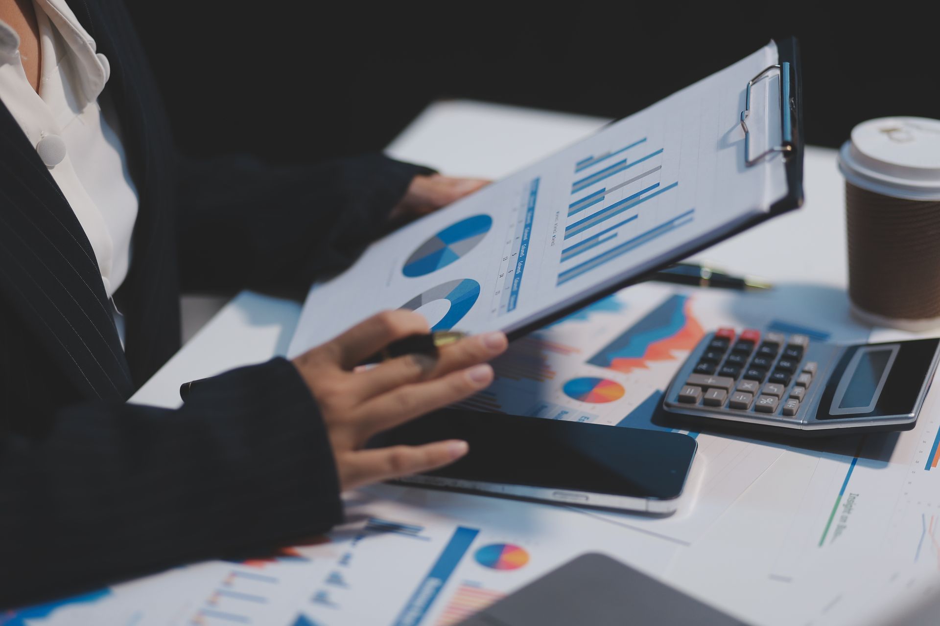 Person reviewing charts on a tablet beside printed graphs, calculator, and coffee cup at a desk