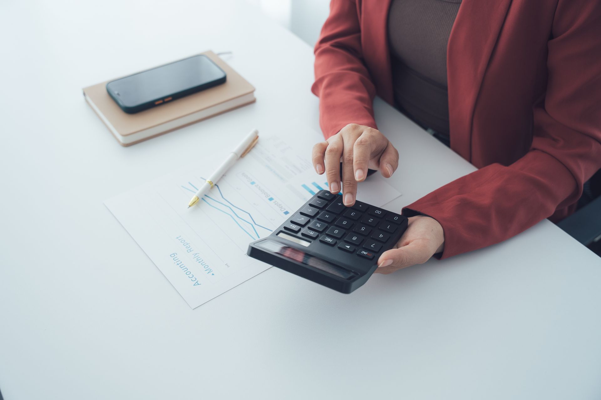 Person in red blazer using a calculator at a white desk with notebook, pencil, and smartphone.