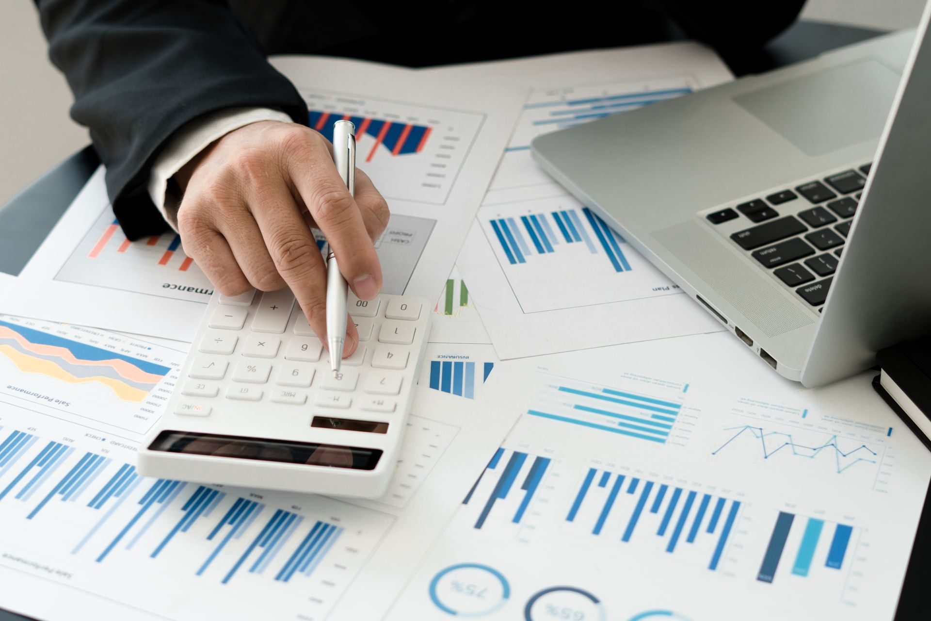 Hand using calculator over financial charts beside a laptop on a desk