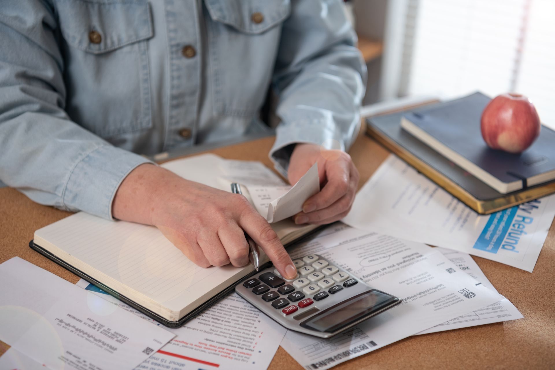 Person using a calculator and notebook at a desk with paperwork and a red apple nearby