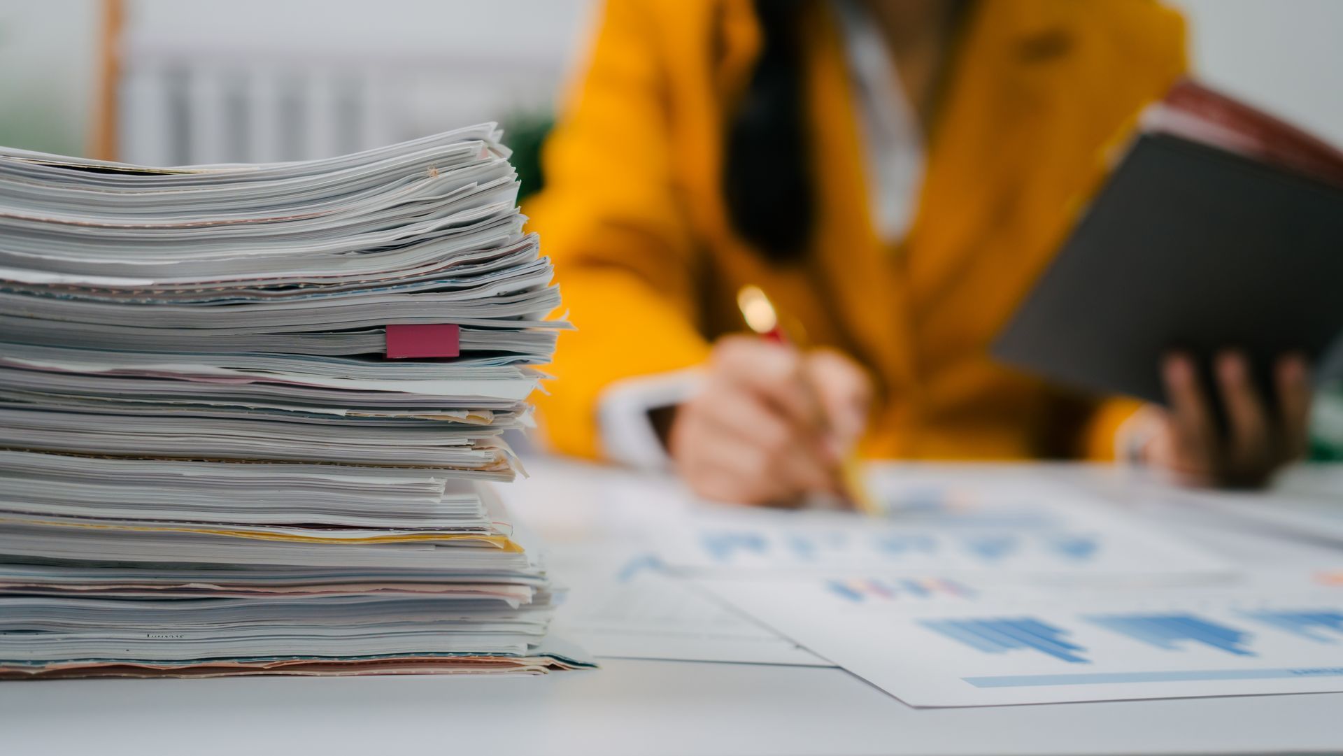 Stack of papers beside a person in a yellow jacket writing on documents at a desk