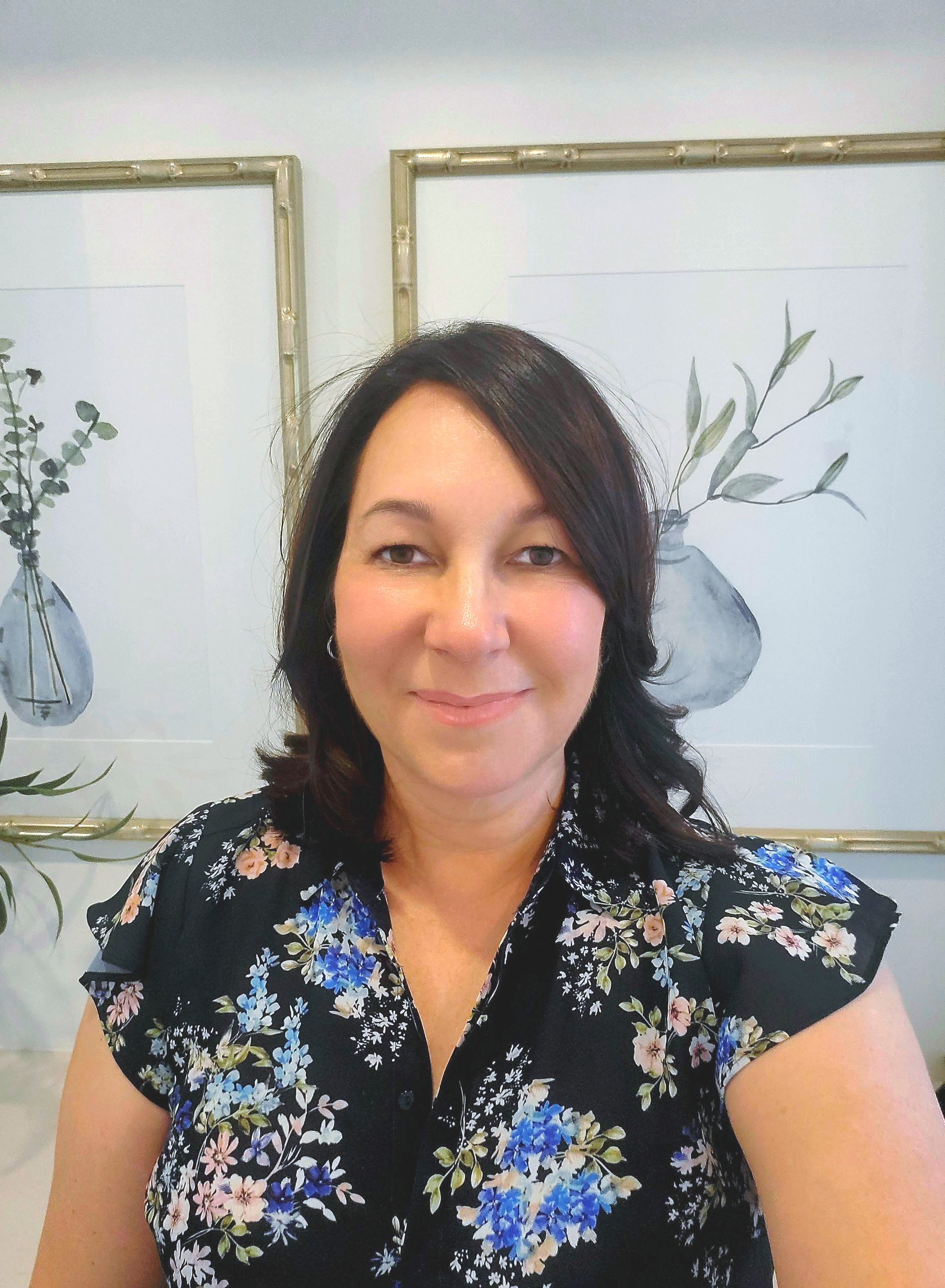 A smiling person with dark hair wears a black floral blouse, positioned before two framed botanical prints on a wall— Jodi Maree Interiors in Dubbo, NSW