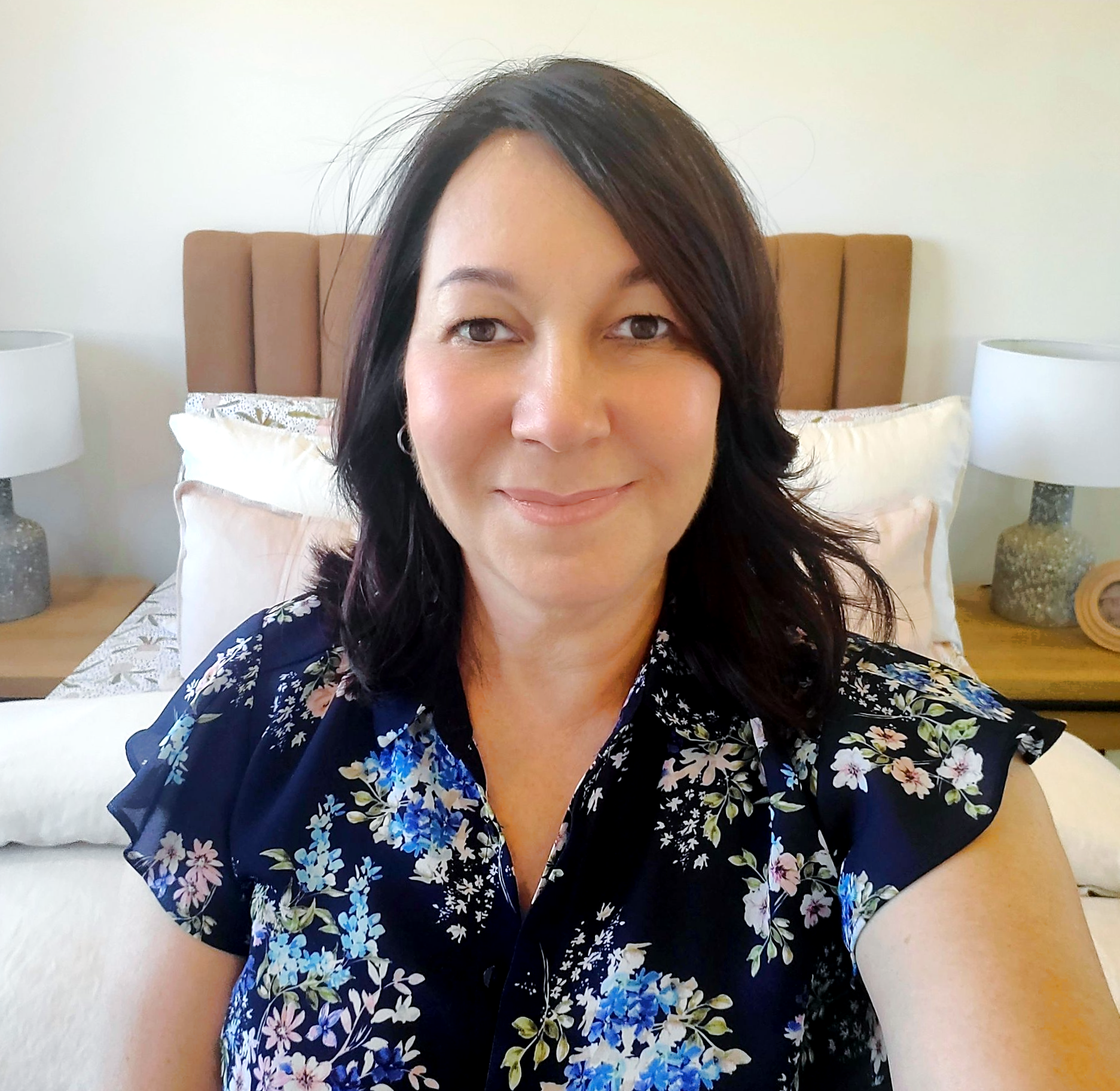 A person with dark hair wearing a navy blue floral top, smiling in front of a bed — Jodi Maree Interiors in Dubbo, NSW