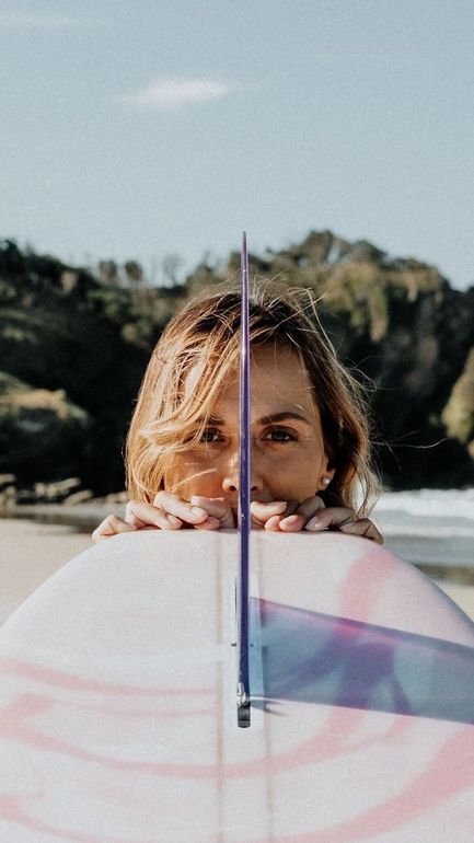 Person peeks over a pastel surfboard on a beach, with rocky cliffs in the background Legs on a surfboard, riding the waves — Holiday Sun Swimwear on the Gold Coast, QLD