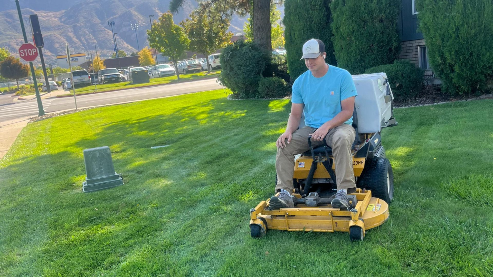 Man on a yellow lawn mower cuts grass in a bright green yard with a road and mountain backdrop.