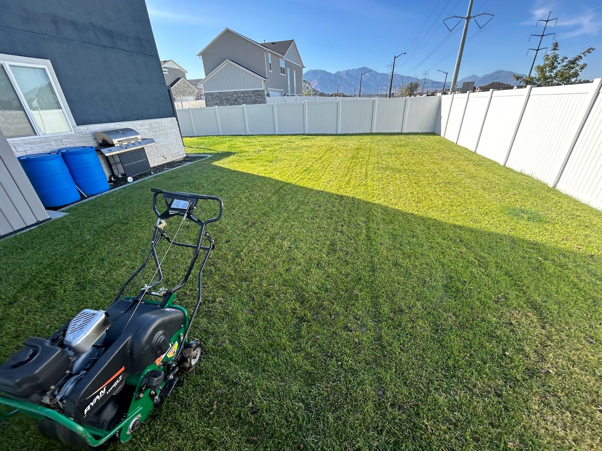 Lawn mower in a green backyard surrounded by a white fence on a sunny day with mountains in the background.
