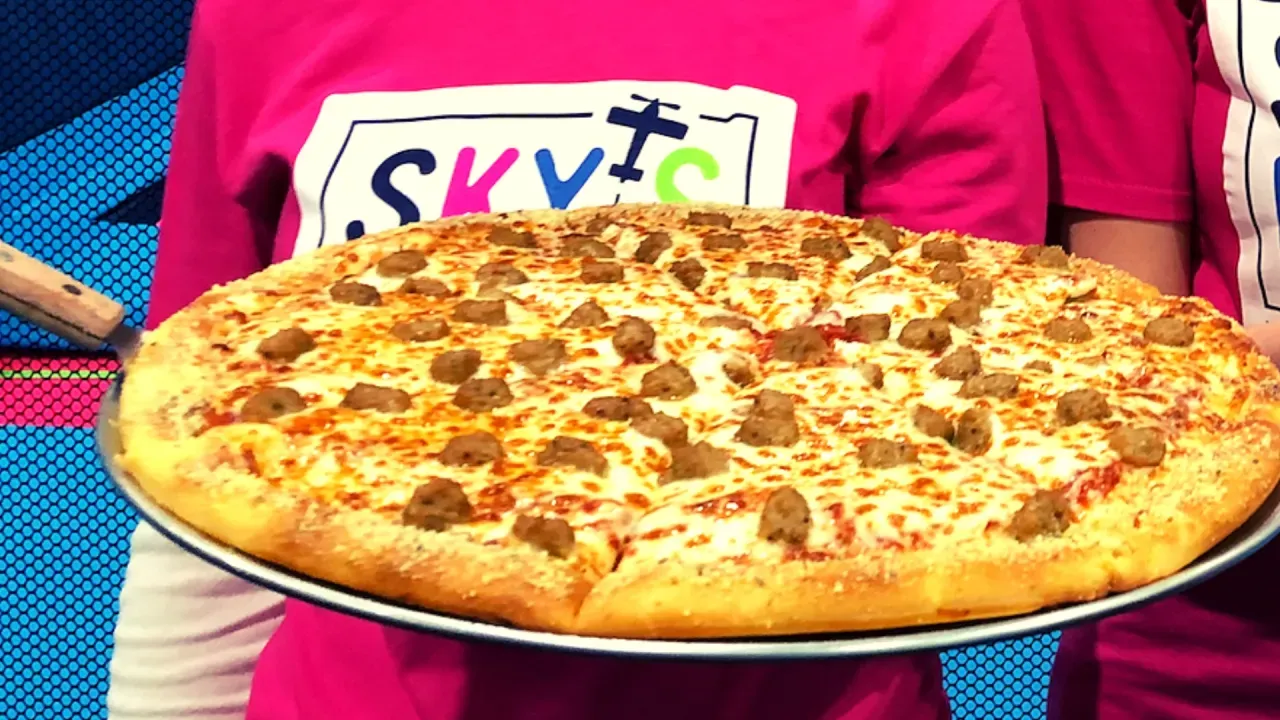 Person holding a large, round pizza with sausage on a metal tray. Pink shirt with logo visible.