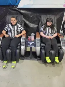 Two people in referee shirts and yellow socks sit in massage chairs with drinks.