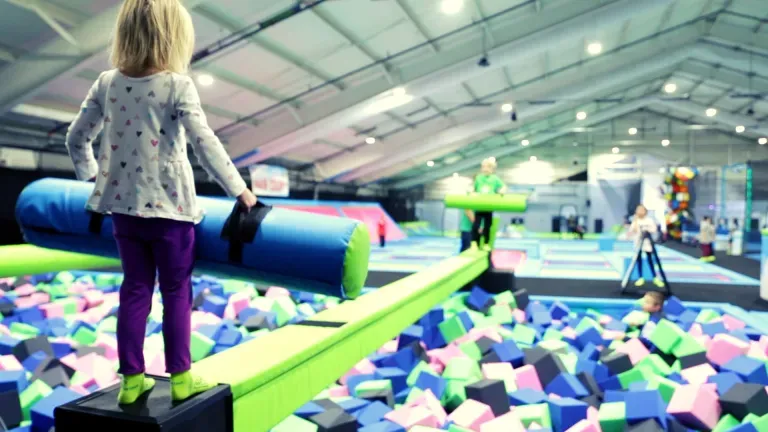 Child on a balance beam, with a foam pit and trampoline park in the background.