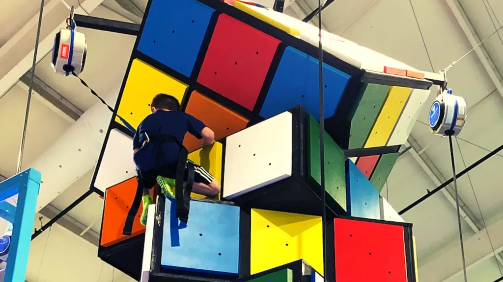 People climbing a large Rubik's Cube structure indoors. Colored blocks, safety harnesses, and a high ceiling.