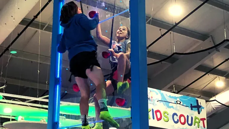 Two people climbing a clear indoor climbing wall with blue lights.