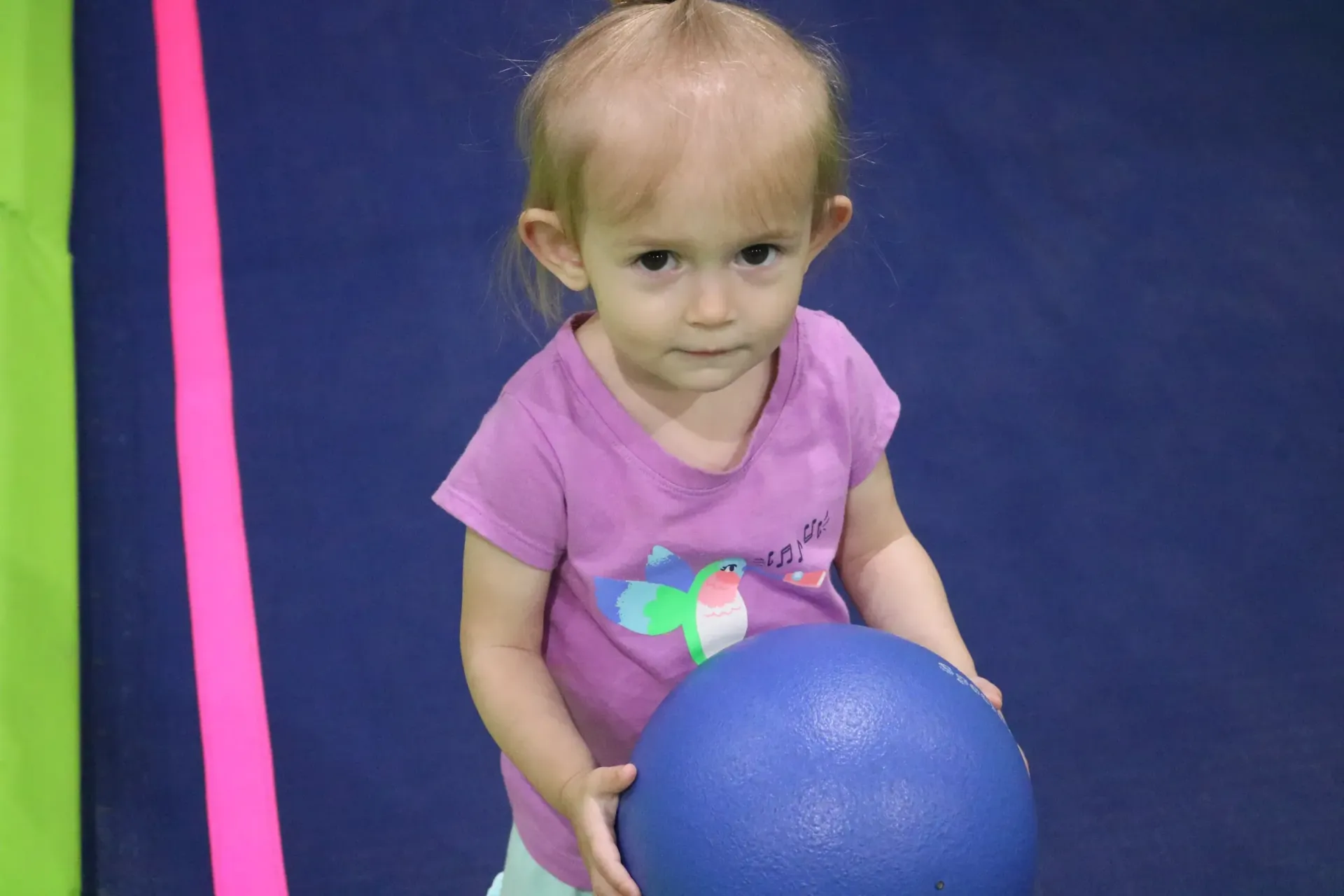 Young child in purple shirt holding a blue ball, looking towards the camera. Background has green, pink, and blue.