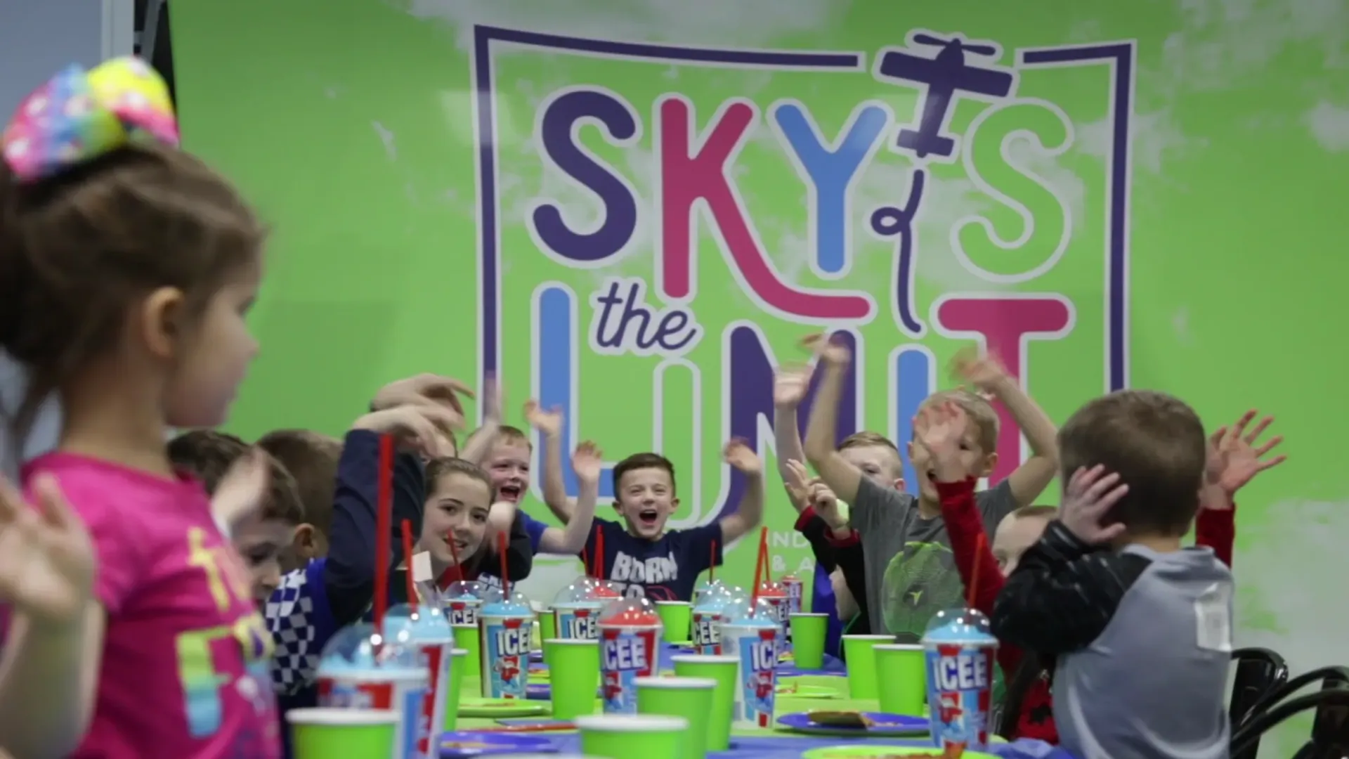 Children at a party, cheering. Green and purple backdrop with 