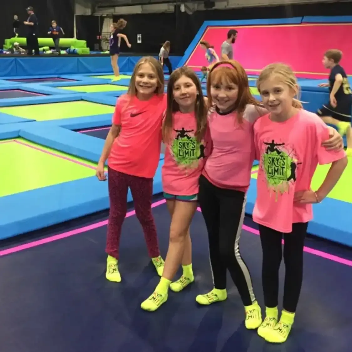 Four girls in pink shirts and yellow socks pose on trampolines in a trampoline park.