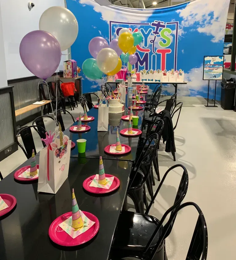 Party table set with pink plates, unicorn hats, and balloons at an indoor event.
