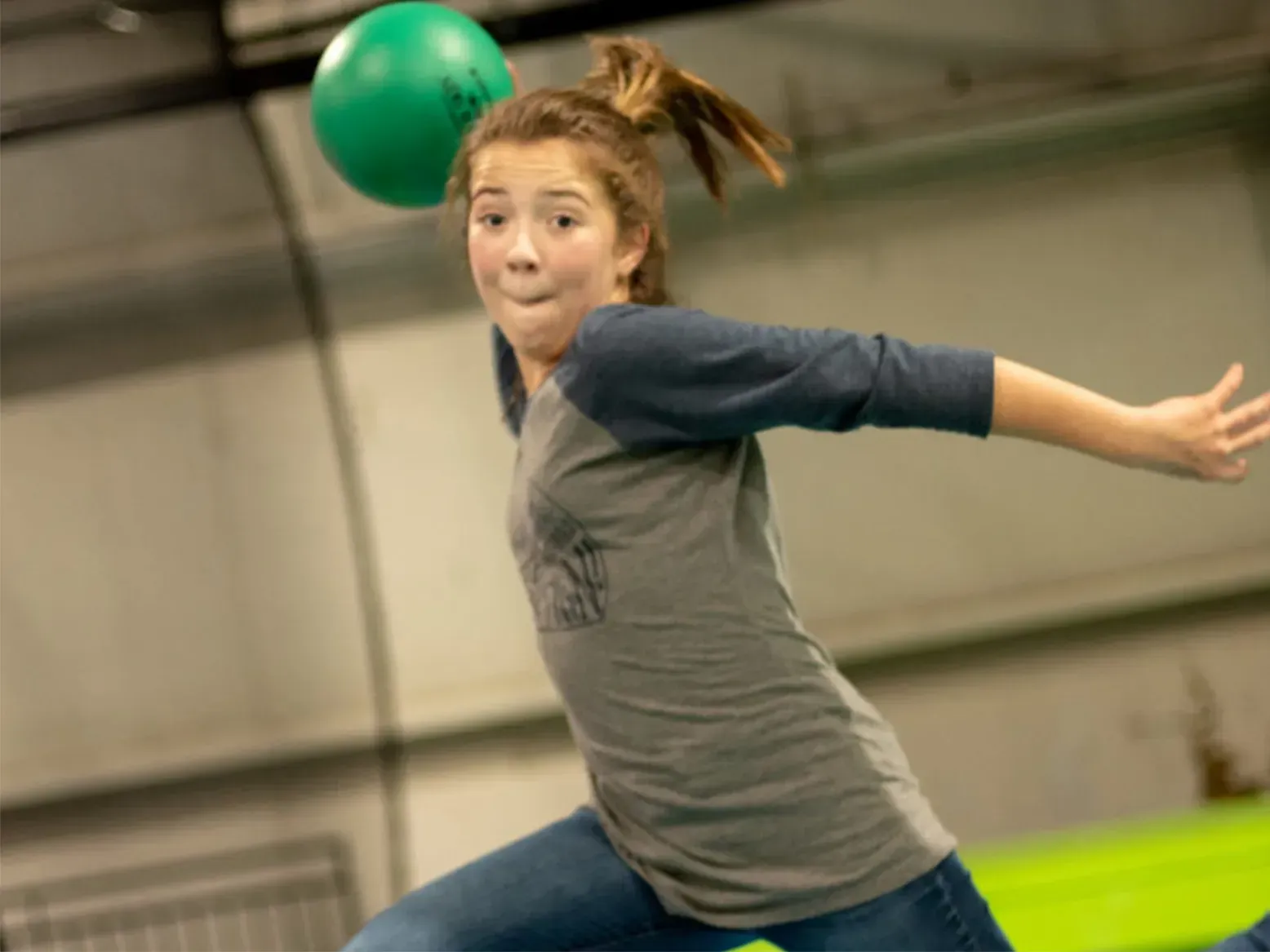 Girl throwing green ball, indoors. Wearing gray shirt, jeans. Focused expression, arm outstretched.
