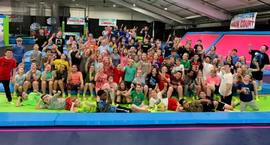 Large group of people on a trampoline, many with arms raised, inside a colorful recreation center.