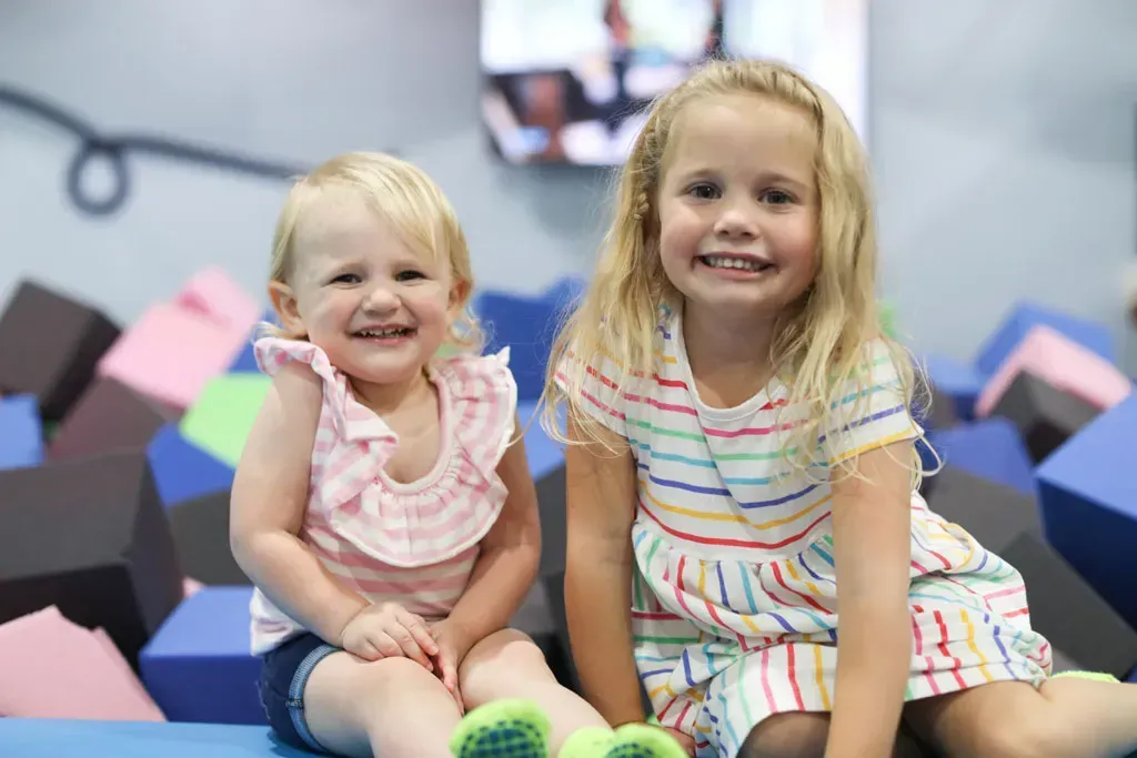 Two smiling young girls sit near colorful foam blocks.