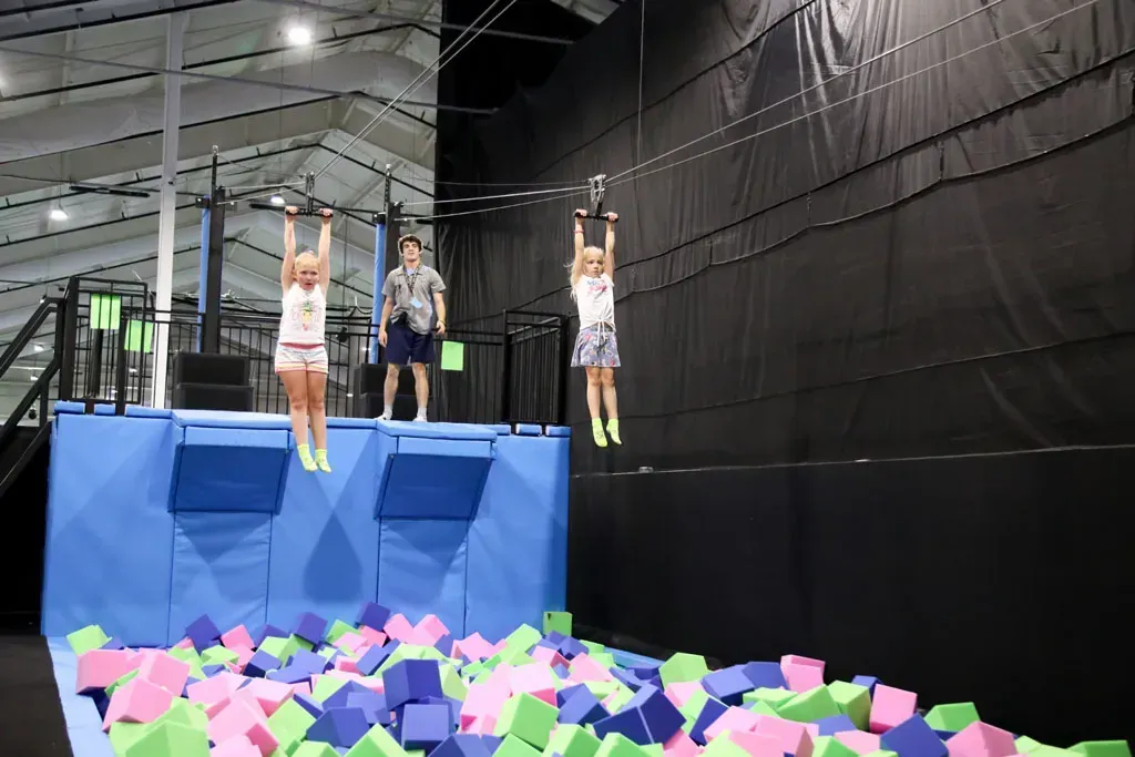 Two children zip-lining over a foam pit, another person observes at an indoor trampoline park.