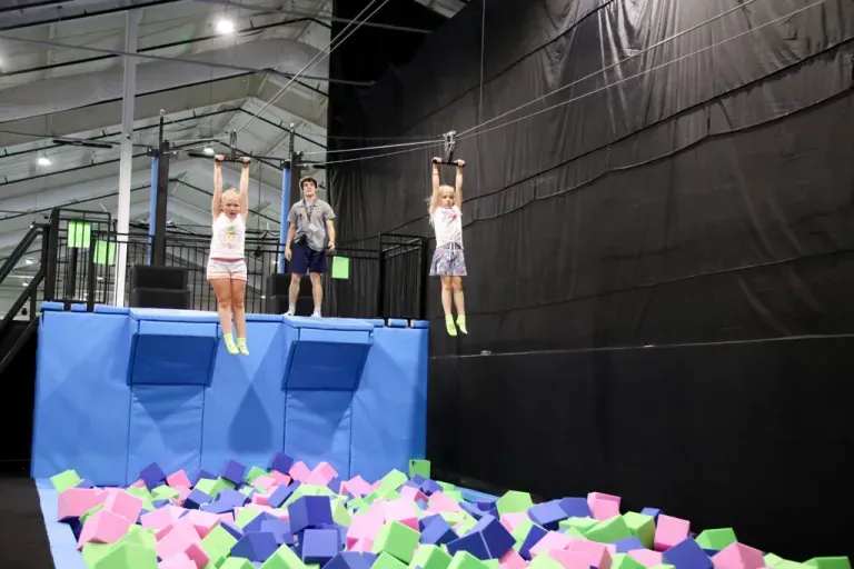 Children on a zip line over foam cubes. Indoor trampoline park setting; a boy watches.