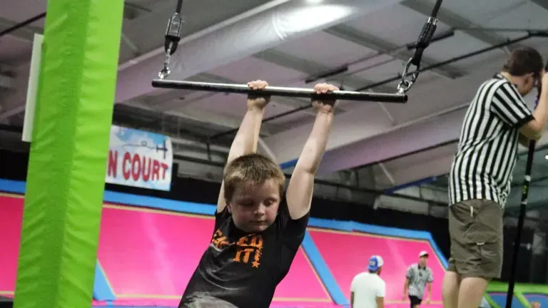 Boy hanging from a bar at an indoor trampoline park. He's wearing a black shirt, and a referee is nearby.