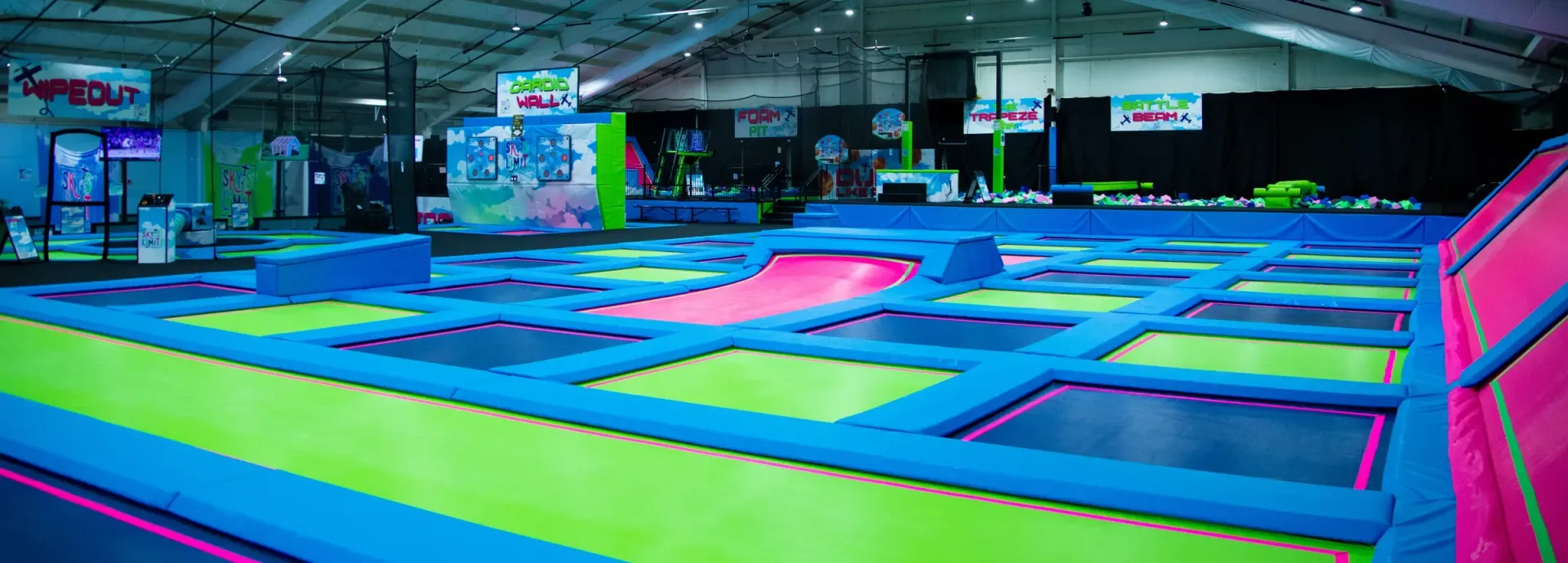Indoor trampoline park with blue, green, and pink surfaces, a ramp, and high ceilings.