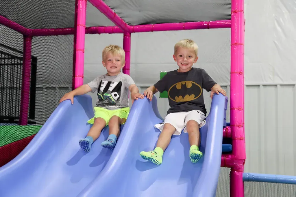 Two young boys on blue slides, smiling, in a colorful indoor play area.