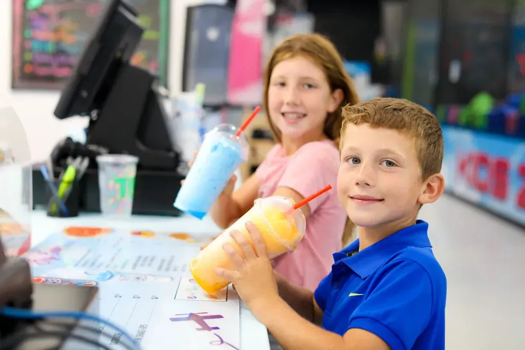 Two children smiling, holding colorful slushies at a counter in a shop.
