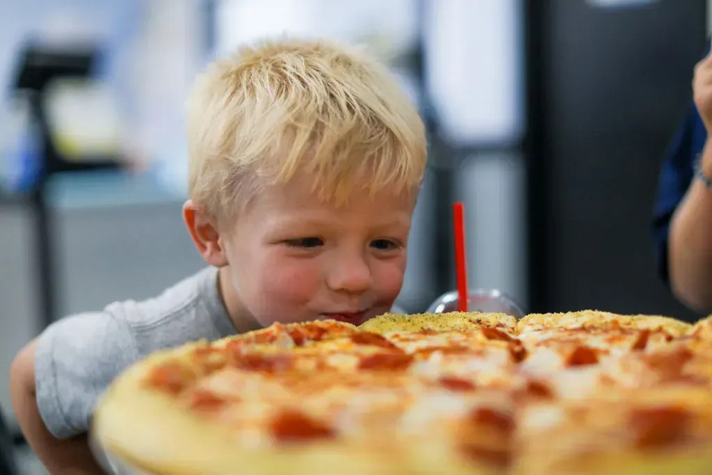 Blond child looks at a large pizza, his face close to the crust, with a drink in the background.