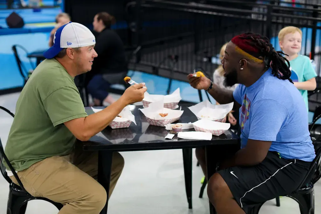 Two men eating at a black table indoors. One wears a green shirt, hat; the other a blue shirt, with red and blonde hair.