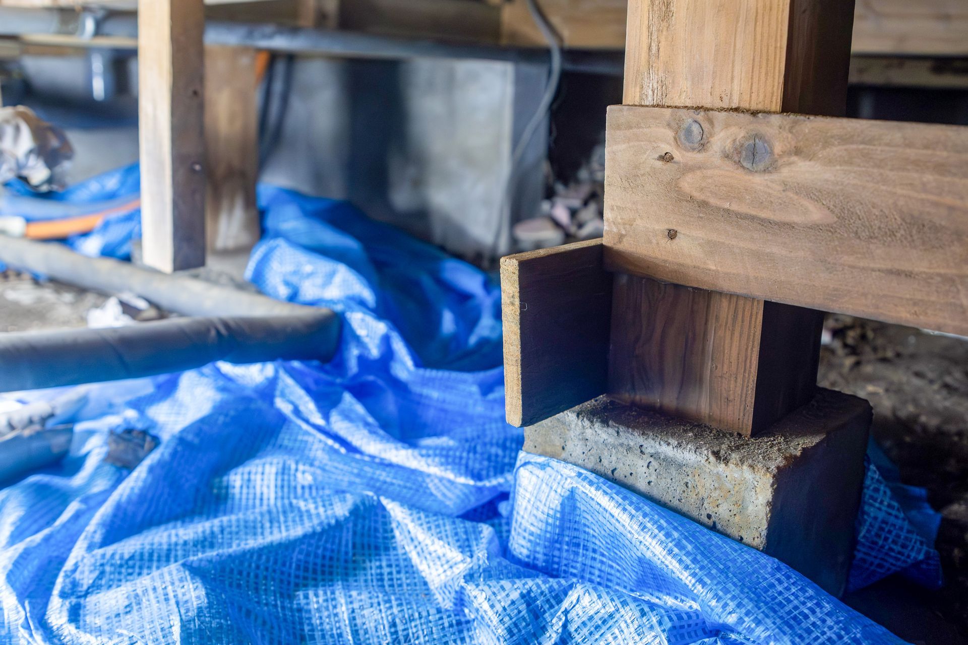 Wooden foundation support covered in blue tarp under a house during repair work. The inspector is checking rebar and layout during concrete foundation work.