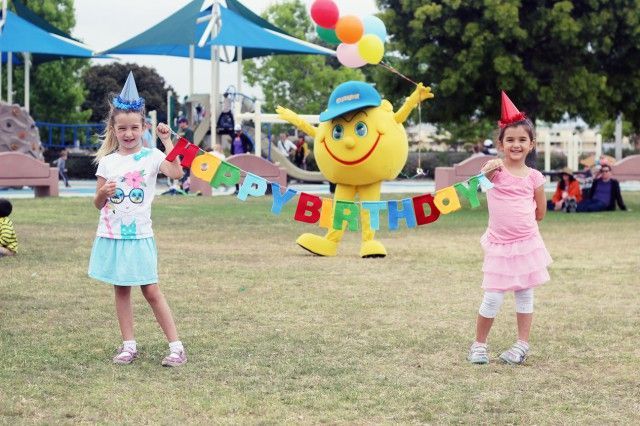 Two little girls are standing in front of a mascot holding balloons and a banner that says happy birthday