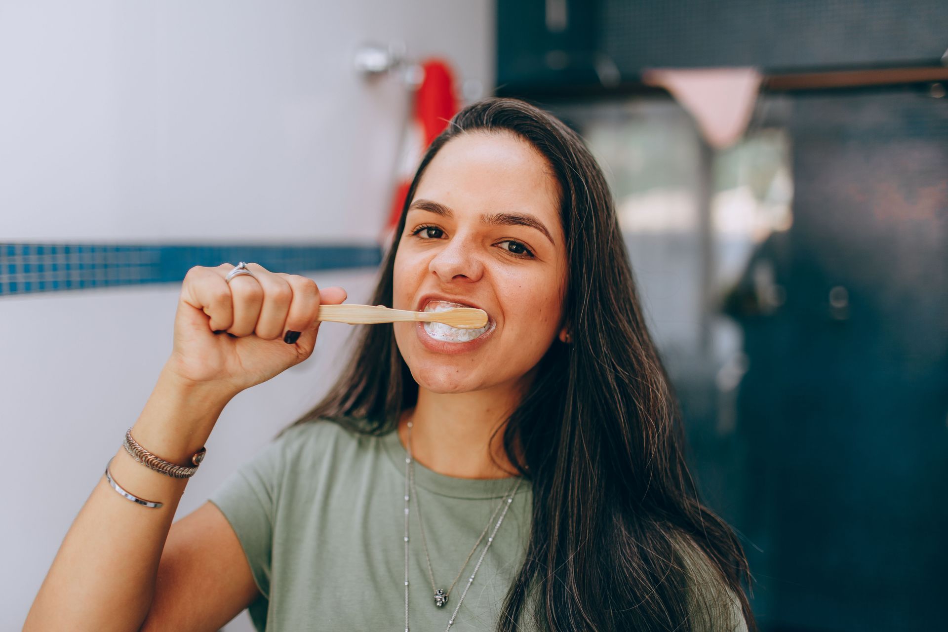 Woman brushing her teeth outdoors with a wooden toothbrush, smiling in a casual green shirt.