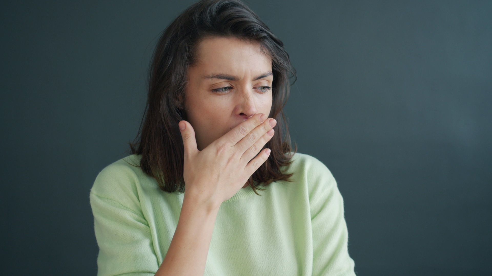 Person in a light green sweater covering their mouth, looking pensive against a dark background.