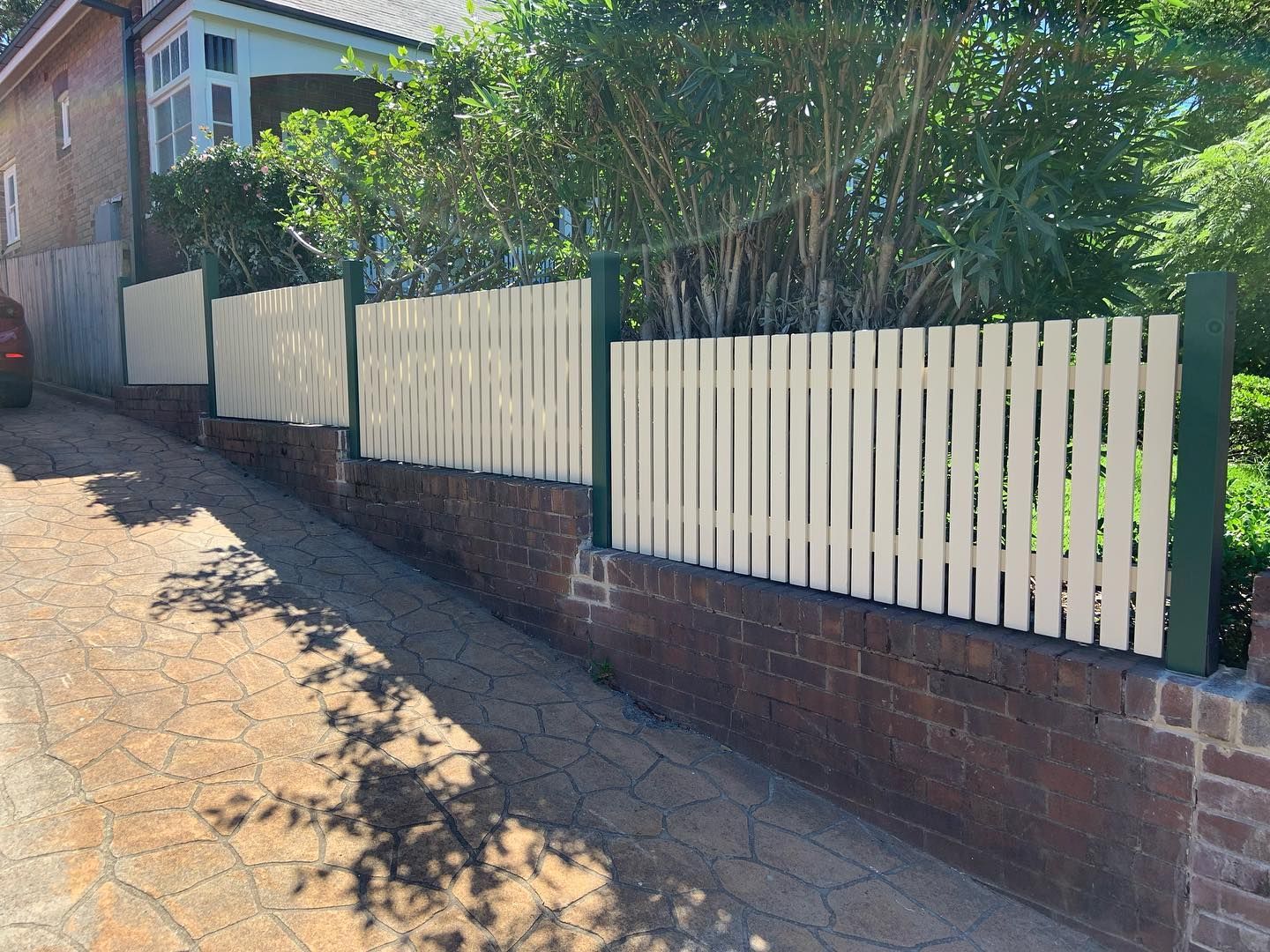 A tan picket fence stands atop a dark brown brick wall along a paved, sloped driveway next to a house.