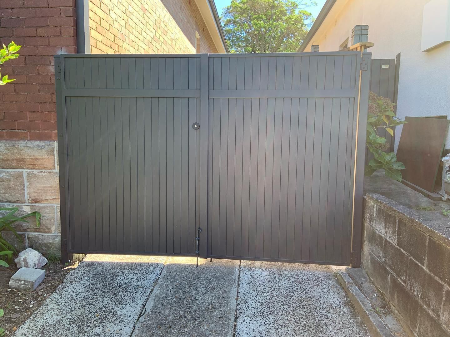 A dark-colored, double-gate fence made of vertical slats installed in a paved residential driveway.