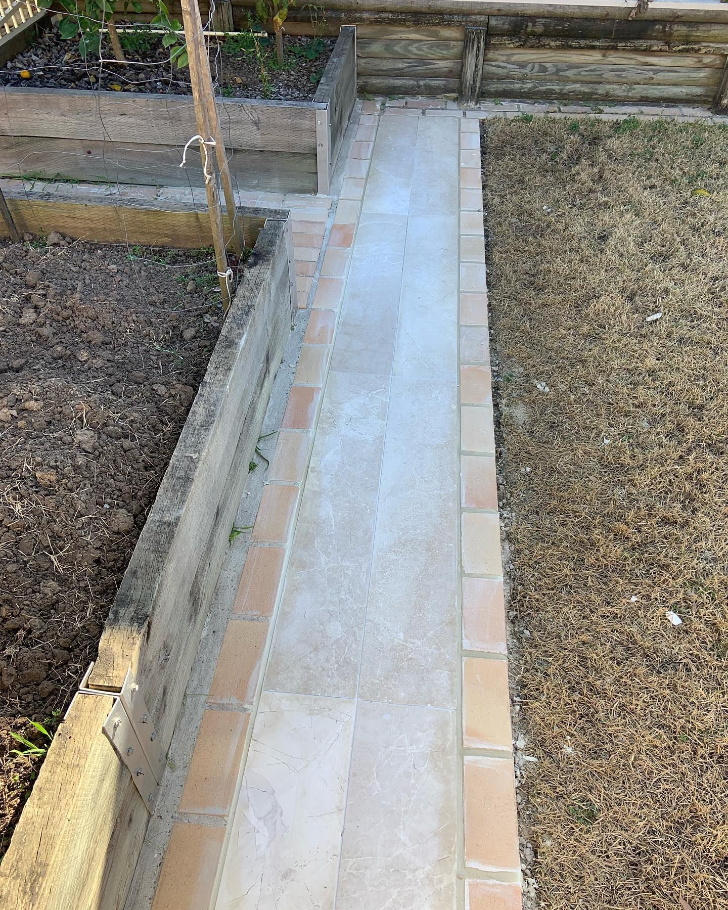A straight paved garden path with reddish-brown brick borders runs between a raised garden bed and a patch of gravel.