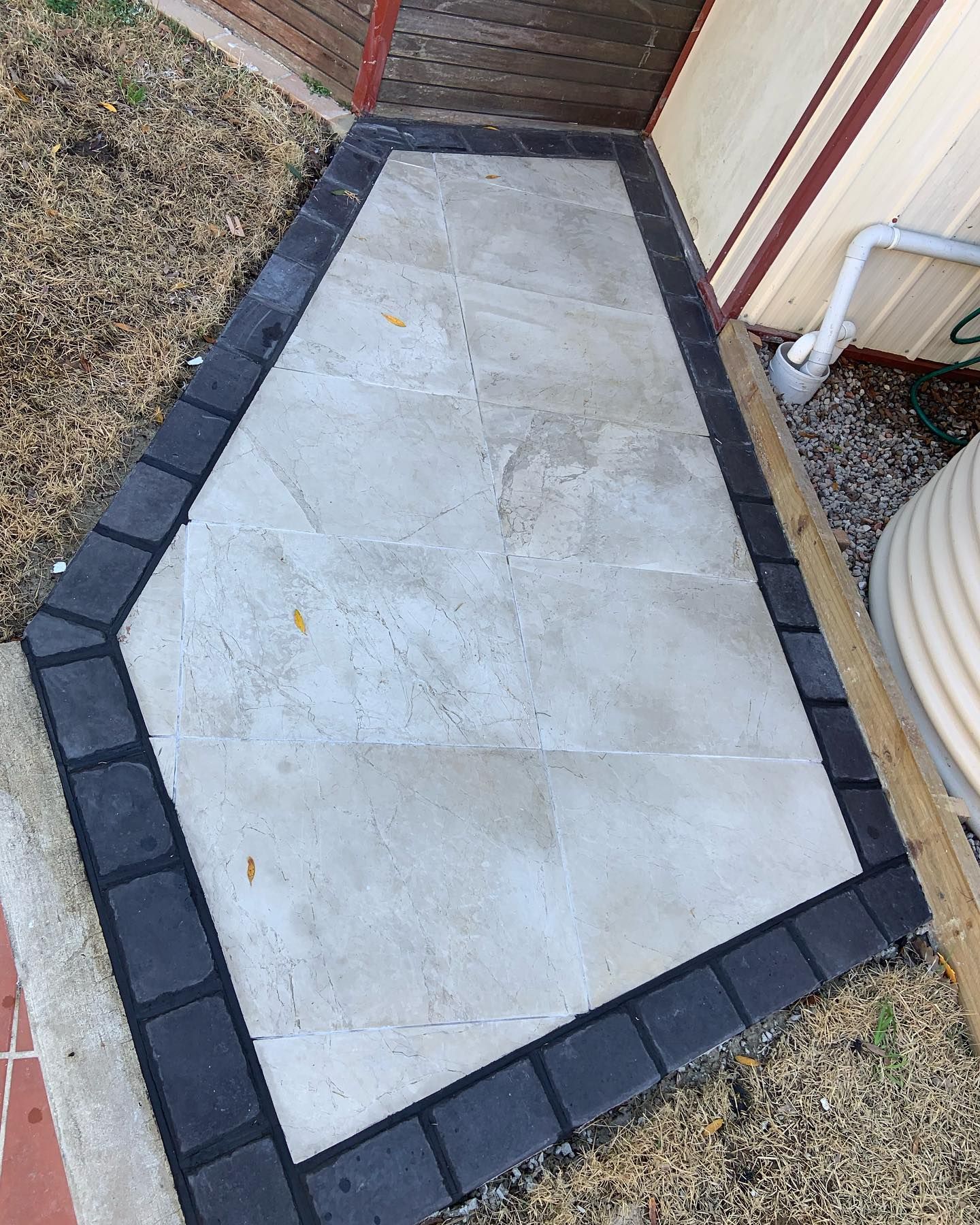 A light-colored stone walkway framed by a border of dark rectangular pavers, set against a wall and landscaped area.