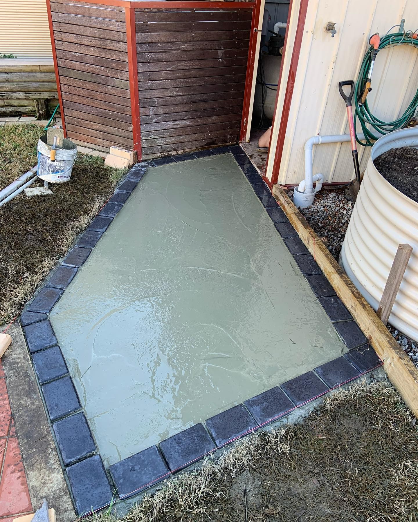 A wet concrete walkway framed with dark charcoal pavers beside a wooden screen and a white corrugated garden bed.