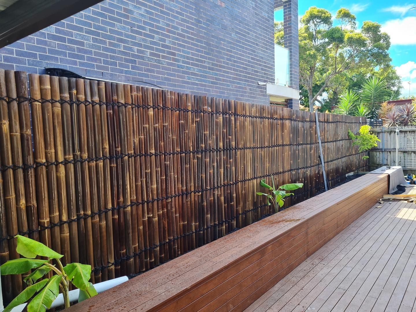 A dark bamboo screen fence stands beside a raised wooden garden bed and decking area against a grey brick wall.