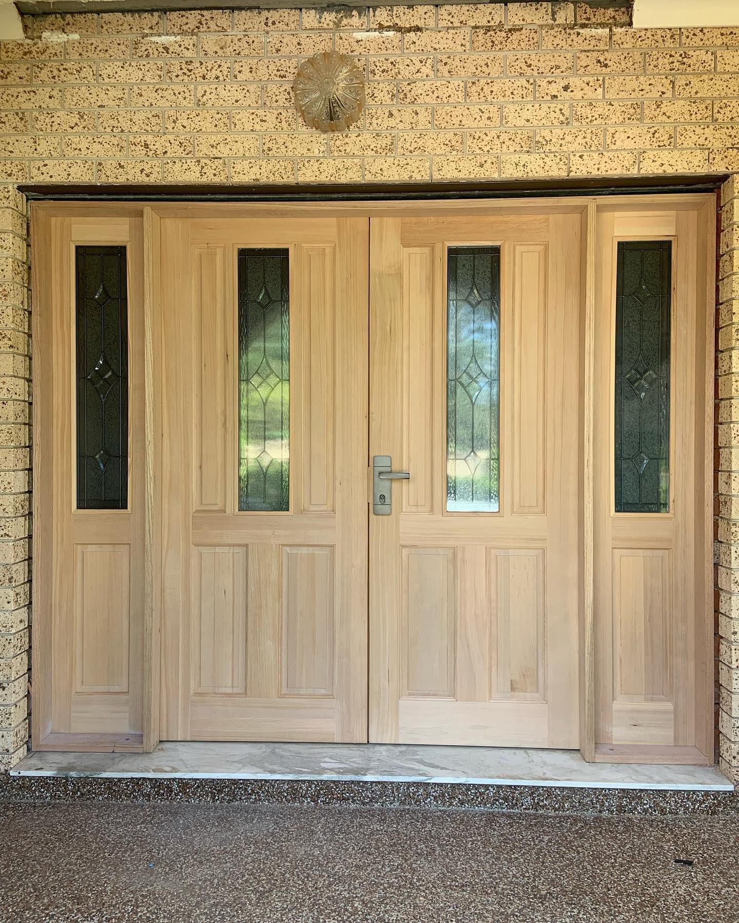 A set of unfinished light wood double entry doors with side panels featuring long glass inserts, set in a stone wall.