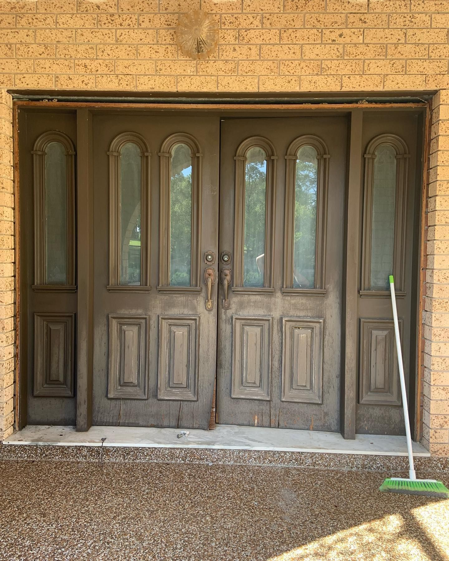 Weathered wooden double front doors with arched glass panels set in a brick wall, broom leaning on the right side