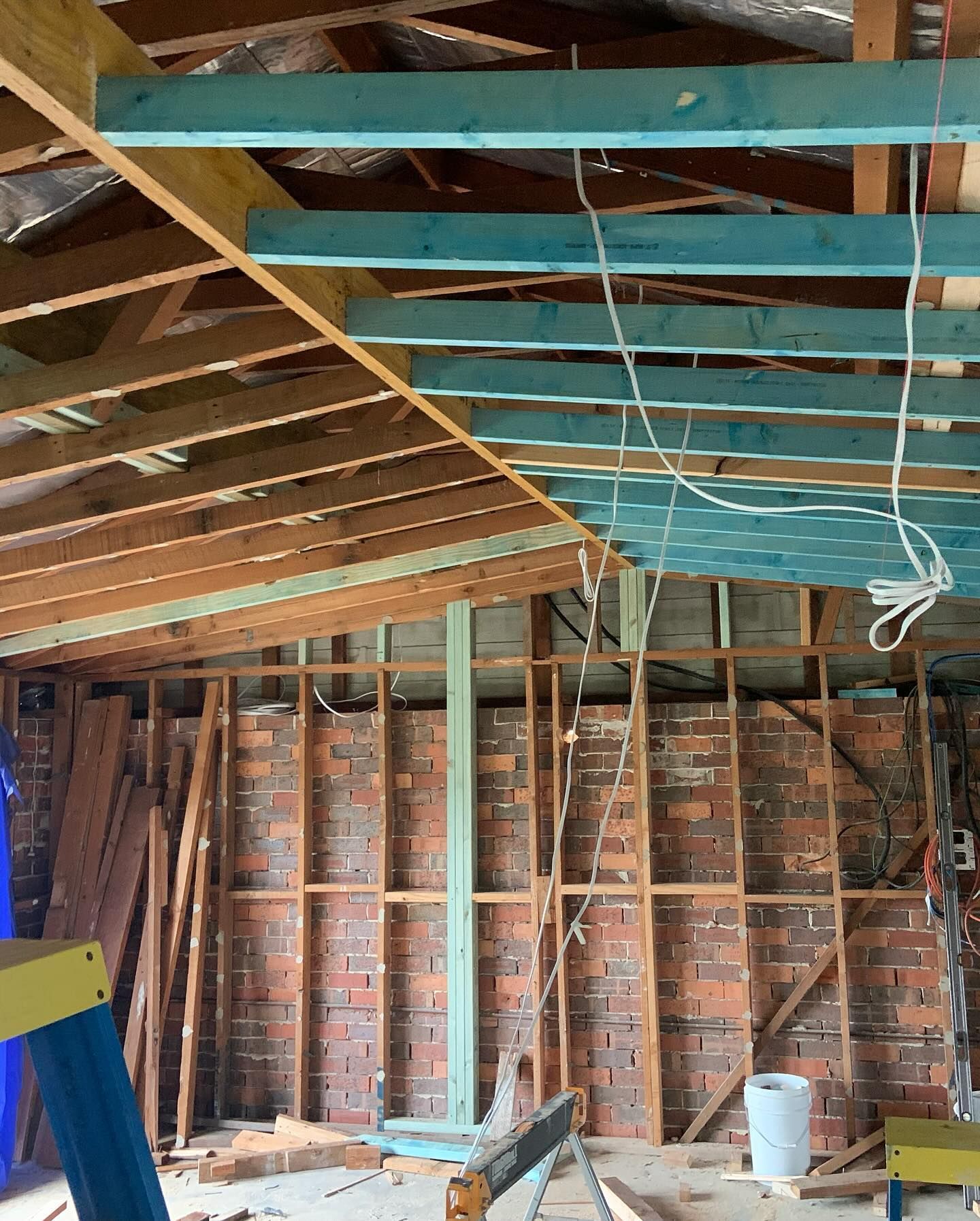 Construction site showing exposed wooden roof rafters, blue ceiling joists, and a partial brick wall frame.