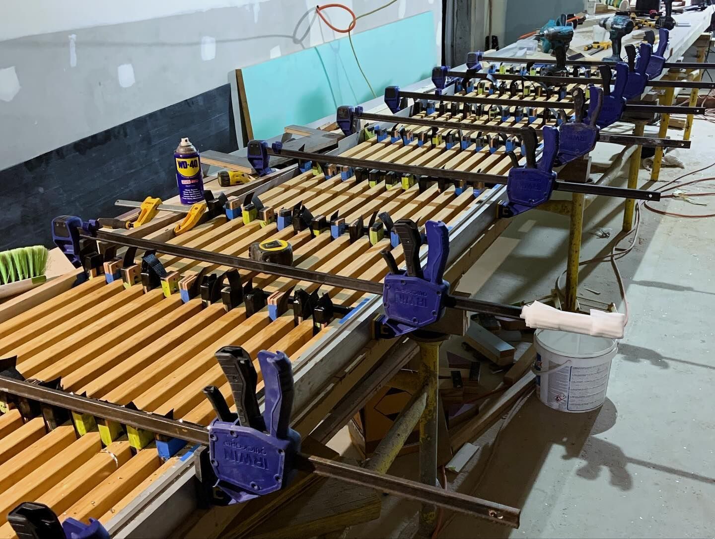Multiple wooden boards are clamped together on a workbench in a workshop, undergoing a gluing process.